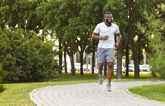 A man wearing a white t-shirt and grey shorts jogs on a paved path through a park, smiling as he listens to music. - Powered by Adobe