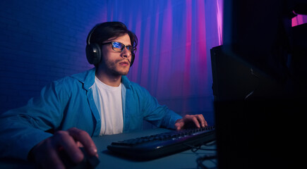 A man wearing a blue shirt and glasses is focused on his computer screen. He is using a keyboard and mouse, and has headphones on. The room is lit with blue and pink neon lights.