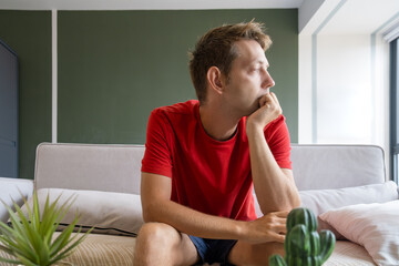 Thoughtful man in a red shirt sits on couch at home and looking in the window. Tired bored male sitting on sofa in modern living room. Stress, depression melancholy and mental health concept