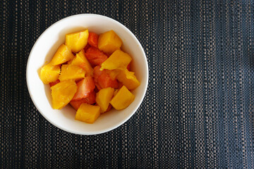 Tropical Fruit Bowl. A white bowl filled with fresh papaya and pineapple chunks placed on a dark textured tablecloth