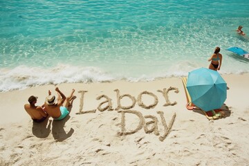 Two individuals sit on a beach with labor day written in the sand, enjoying the coastal scenery. The setting suggests a leisurely and funfilled time by the water
