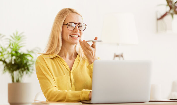 A woman sits at a desk, wearing glasses and a yellow shirt, smiling while talking to her phone, likely using a voice assistant, while working on a laptop. - Powered by Adobe