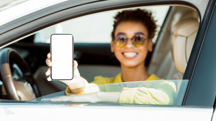 African American woman is sitting in the drivers seat of a car with the window down. She is smiling and holding a smartphone with a blank screen towards the camera.