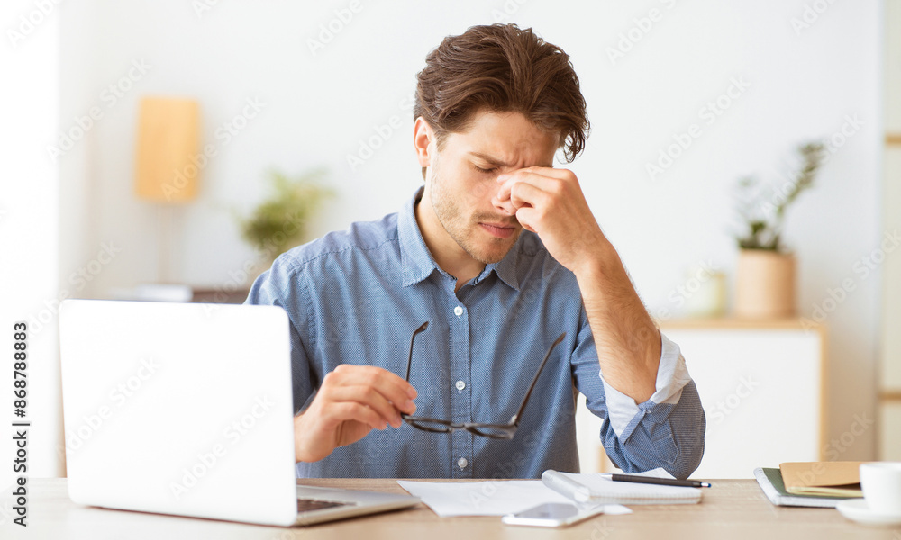 Poster a man sits at a desk, rubbing his eyes in exhaustion. he is working on a laptop and appears to be ex