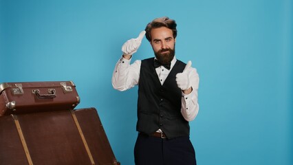 Hotel concierge shows thumbs up in studio, acting positive and optimistic against blue background. Joyful doorman does like and agreement symbol of acceptance, wearing formal attire.