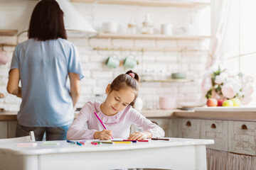 A young girl sits at a white kitchen table, diligently coloring with a purple crayon, focused on...