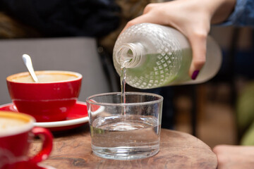 Close-up of a womans hand pouring water into a glass from a glass bottle in a cafe, on a wooden table with coffee cups