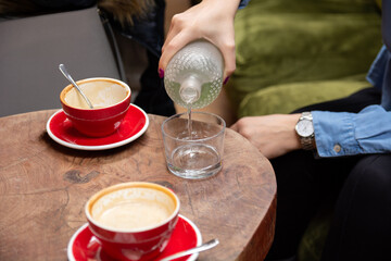 Close-up of a womans hand pouring water from a glass bottle into a glass in a cafe, on a retro wooden table with two red coffee cups