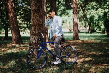 Obraz premium A young man in casual attire stands next to his blue bicycle in a lush green park, speaking on his mobile phone under the shade of trees.
