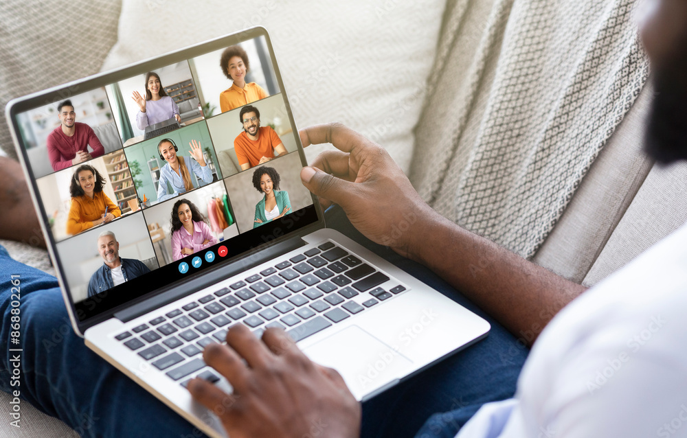 Poster black man sits on a couch, using a laptop to participate in a video conference call with multiple ot