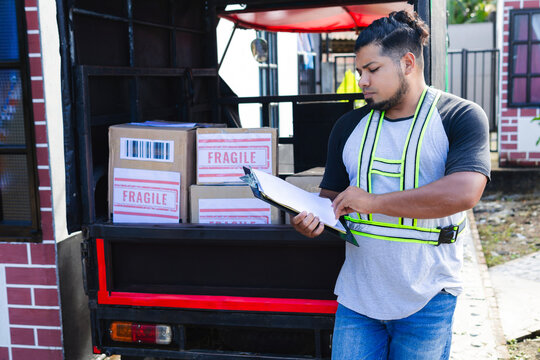 Colombian Latino man working at a national shipping company, checking notes for upcoming destinations. The vehicle, a motorized tricycle, is filled with orders