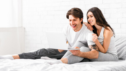A happy couple relaxes in bed, the man is holding a laptop while the woman holds a mug of coffee and smiles. The couple is enjoying their time together in a cozy setting, copy space