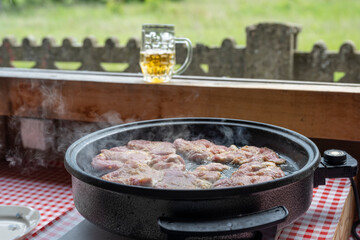 Frying or stewing pork, beef, veal in a special pan. Summer kitchen in the countryside. In the background you can see a mug of beer and a beautiful view of the meadow and the forest.