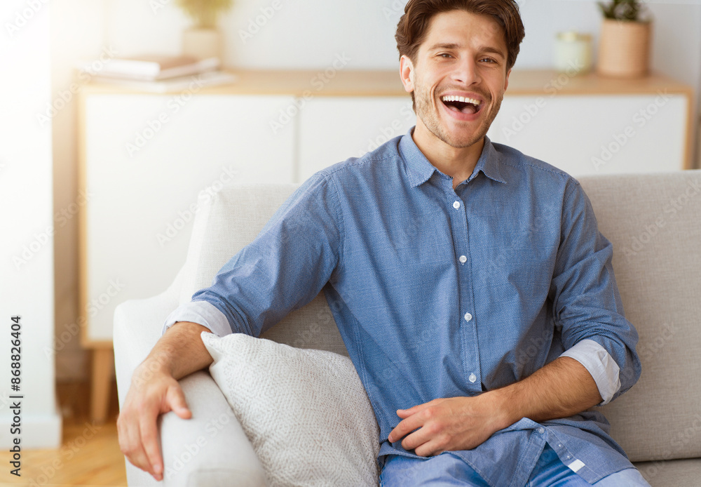 Poster a man in a blue button-down shirt sits on a white couch and laughs. he is relaxed and looks happy.