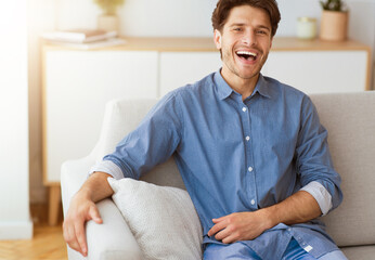 A man in a blue button-down shirt sits on a white couch and laughs. He is relaxed and looks happy.