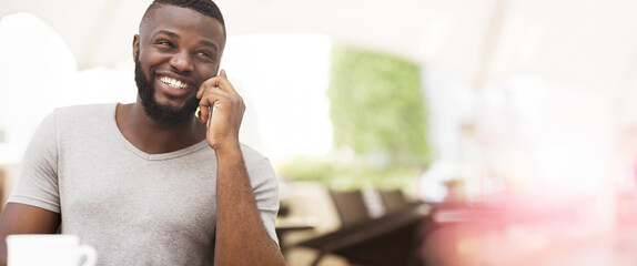 A man with a beard smiles while talking on the phone, sitting indoors with a blurred background.