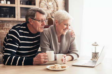 A senior couple is sitting in their kitchen, enjoying coffee and conversation while using a laptop. The woman is smiling at the screen, while the man is looking at her