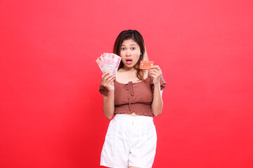 Shocked indonesia woman holding rupiah money and debit credit card wearing brown blouse top with red background. for transaction, technology and advertising concepts