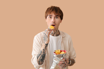 Shocked young tattooed man eating potato chips on beige background