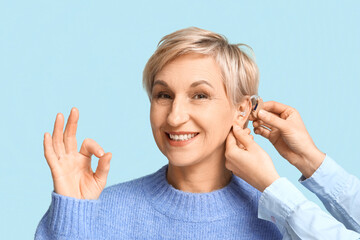 Obraz premium Young woman putting hearing aid in mature woman's ear showing ok gesture on blue background
