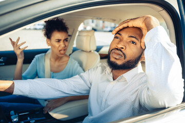 Black man driving a car appears stressed and frustrated as a woman in the passenger seat yells at him. He has his hand on his forehead as he stares forward while she gestures with her hand raised.