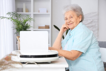Fototapeta premium Senior woman with laptop and robot vacuum cleaner at home
