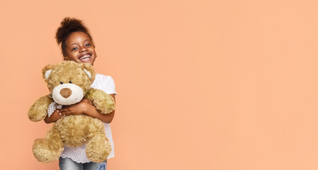 A little black girl with dark hair is smiling brightly as she holds a large, brown teddy bear in...