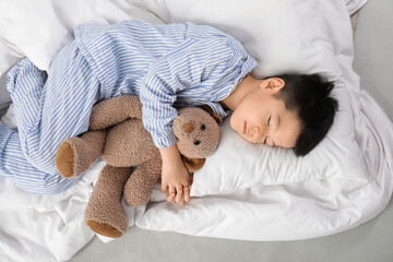 Cute little Asian boy in pajamas with toy bear, pillow and blanket sleeping on white background