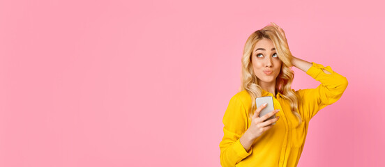 A woman wearing a yellow shirt is holding a smartphone in one hand while using the other hand to adjust her hair. She is standing in front of a pink background and looking at copy space