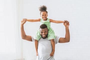 A joyful black father is giving his young daughter a piggyback ride indoors. The girl is wearing a...