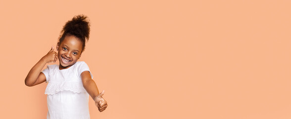 A little black girl with dark brown hair, wearing a white shirt, smiles at the camera while making...