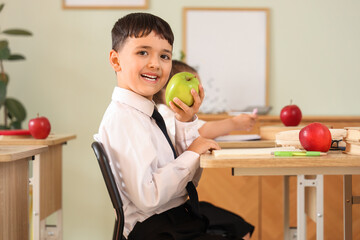 Little schoolboy with apple sitting at desk in classroom
