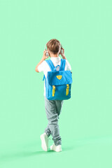 Little schoolboy in headphones with backpack on green background, back view