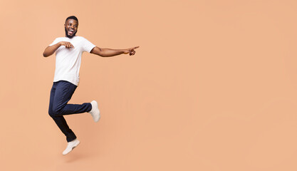A happy African American young man is jumping in the air while pointing to the right with a big smile on his face. He is wearing a white t-shirt and dark blue jeans, panorama with copy space