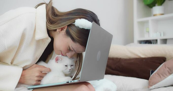 Young woman working on a laptop with a white cat rests on her lap, showcasing a cozy and relaxed home office setup and remote work lifestyle.