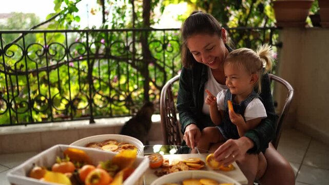 Little daughter sits laughing in the arms of her mother cutting persimmons at a table on the balcony. High quality 4k footage