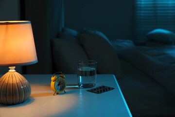Insomnia treatment. Glass of water, pills and alarm clock on bedside table in bedroom at night