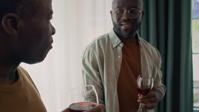 Medium close-up shot of middle-aged African American man and adult son standing together by window at home party, tasting red homemade wine, drinking from glasses and discussing bouquet