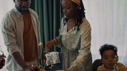 Medium tilt-up shot of young African American woman with braided hair, in long dress and husband putting hot main course meals on table, arranging glasses and plates, while preparing for family brunch