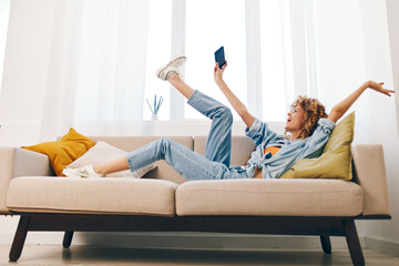 Happy young woman sitting on the cozy sofa at home, holding her smartphone and smiling while chatting online and playing games in the living room.