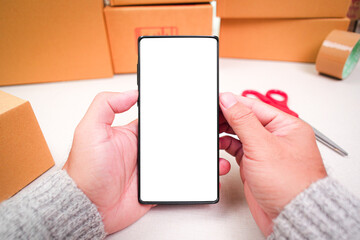 Goods delivery concept , mobile phone with blank white screen in a human hand and cardboard boxes on desk for product packing , copy space for text