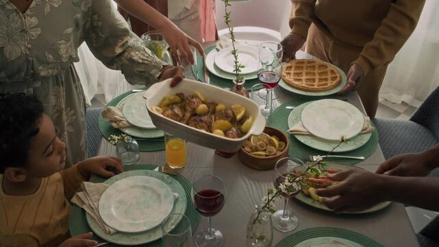 Top shot of African American multigenerational family laying table for Sunday lunch while visiting grandfather, bringing bowls with roast vegetables, chicken, salad and pie, before sitting down to eat