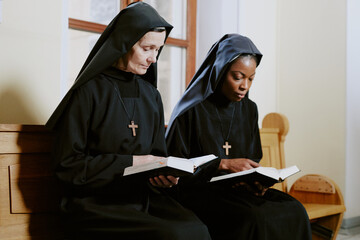Two ethnically diverse nuns sitting on pew in Catholic church and reading prayer books