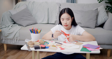 Portrait of Young Asian woman sitting on the floor use scissors to cut paper hearts into stickers to make greeting cards. Holiday hobbies, Handmade, DIY card