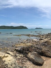 A tranquil coastal view featuring rocky shorelines and calm, turquoise waters. The sky is clear with some clouds, and green islands can be seen in the background. The foreground is dotted with barnacl