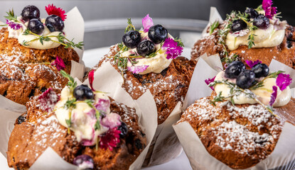 Several muffins on a plate, covered with white icing, blueberries and flowers