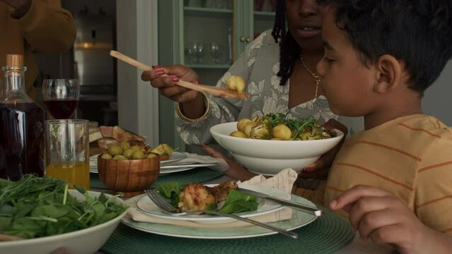 Medium pan shot of smiling African American mother serving roasted potatoes to 6-year-old son at family dinner, grandfather sitting down at table, while celebrating national holiday at home - Powered by Adobe