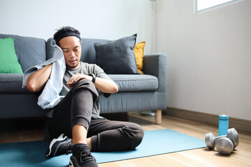 Sporty man sits and looks at smartwatch while wiping sweats after doing exercise at home