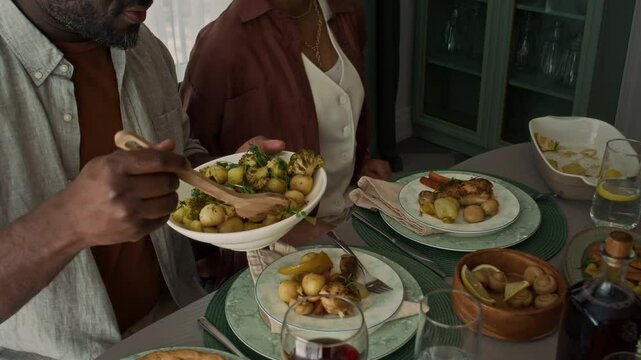 Medium high-angle shot of young African American woman passing bowl of roast potatoes across table to husband during family feast, man serving chicken and vegetables onto plate with spoon