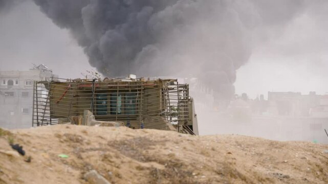IDF Caterpillar D9 armoured bulldozer passing through destroyed streets with bombed buildings and massive fire smoke in background in war between Israel and Hamas